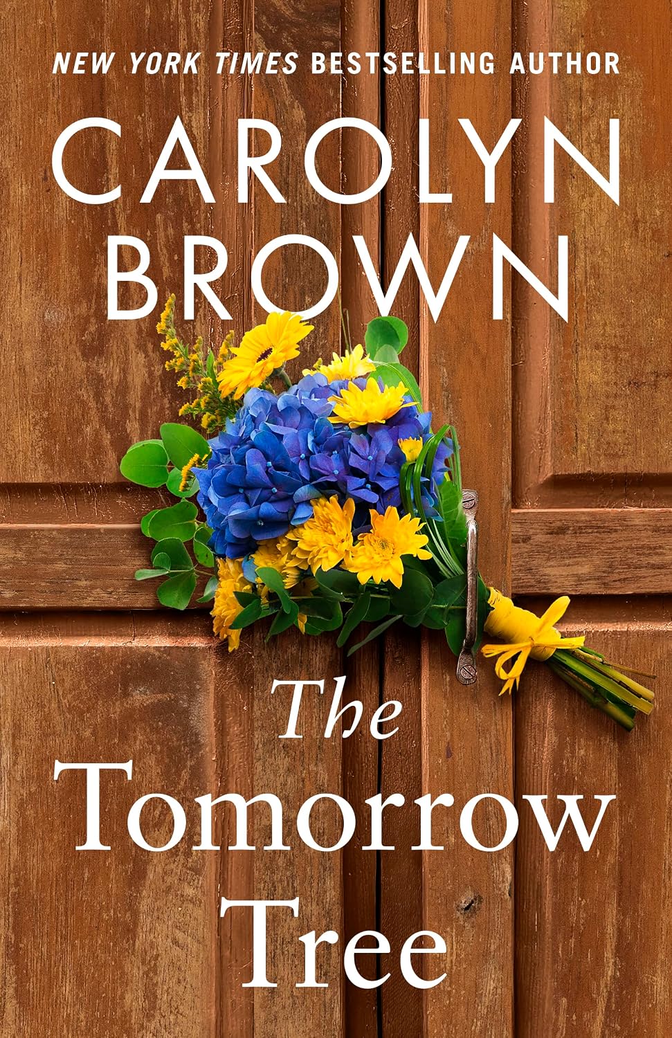 The Tomorrow Tree" by Carolyn Brown. A bouquet of blue hydrangeas and yellow daisies hangs from a handle on a weathered wooden door, with the title in white script below and the author's name at the top.