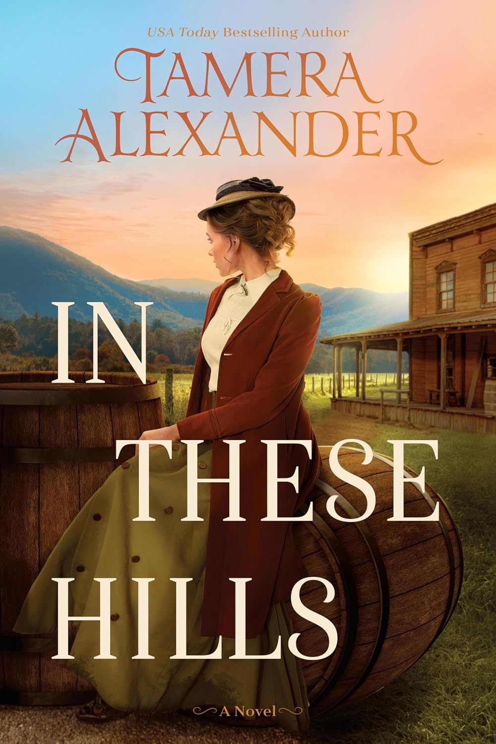 Book cover for "In These Hills" by Tamera Alexander, showing a woman in Victorian-era clothing seated by wooden barrels with mountains and a farmhouse in the background at sunset.