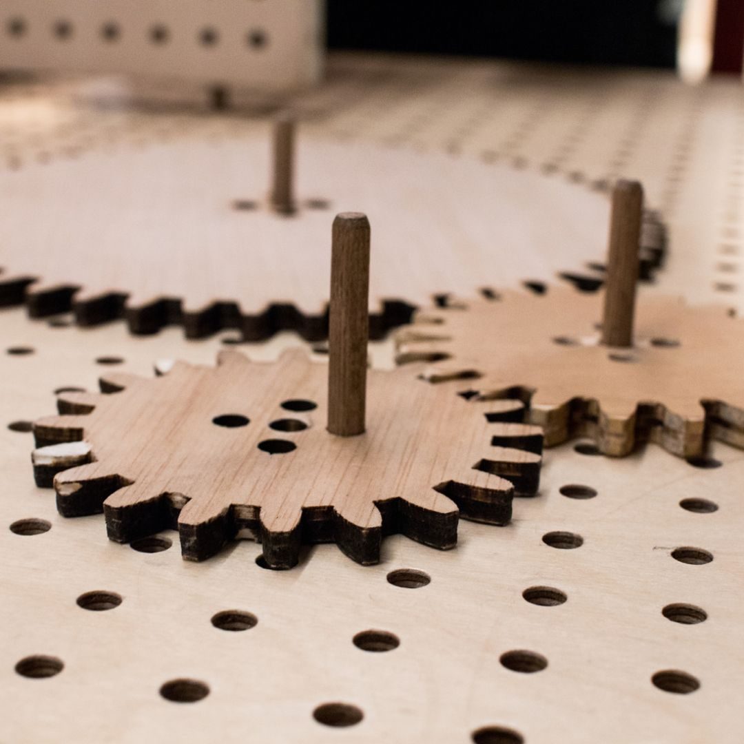 Close-up photo of laser-cut plywood gears with charred edges, each mounted on a wooden dowel peg, sitting atop a pegboard-style surface with rows of drilled holes.